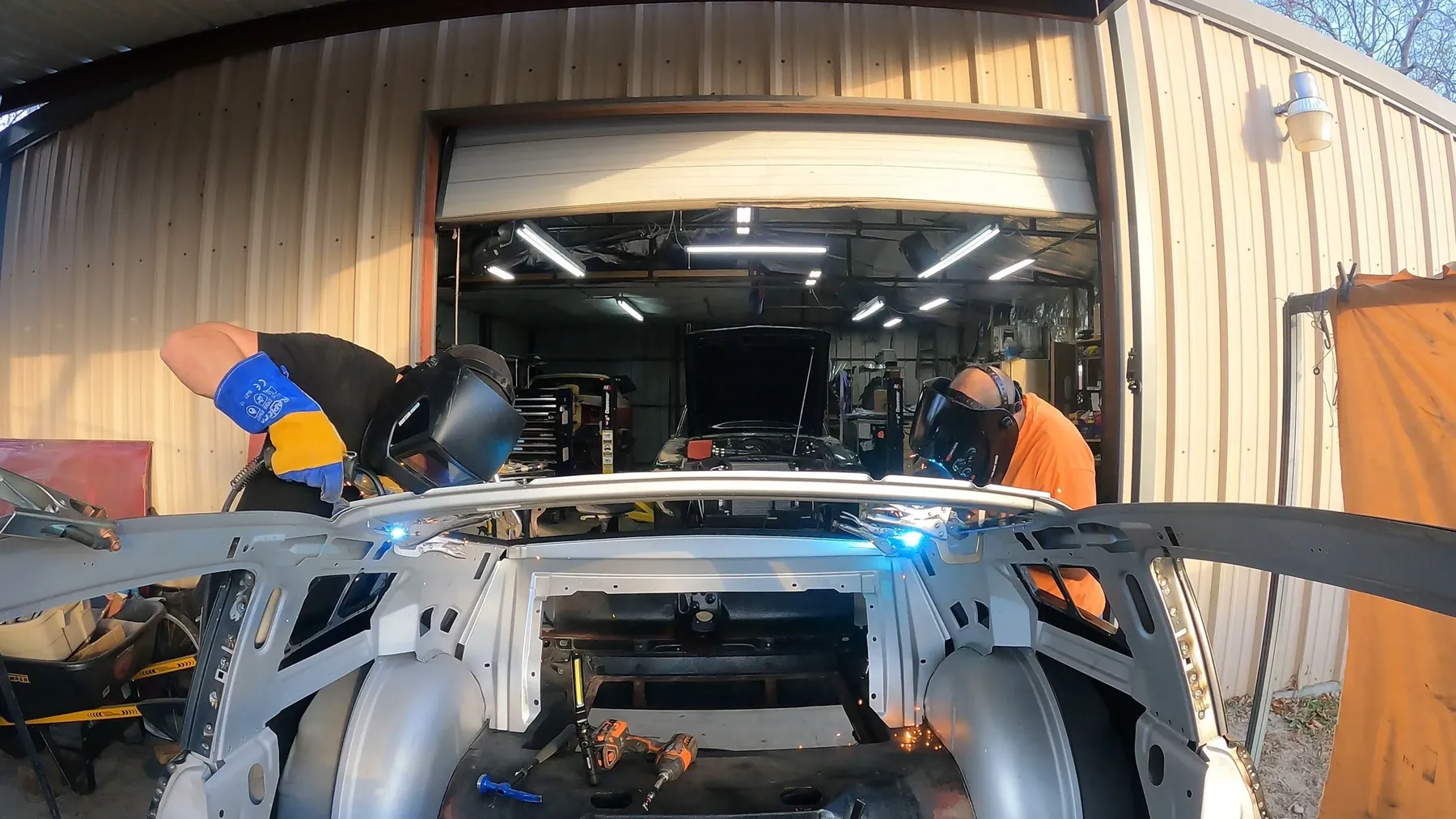 Two builders welding at the front of a Mustang shell under shop lights.